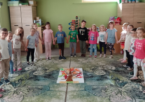 Dzieci stoją w półkolu przy plakacie drzewa z dłońmi